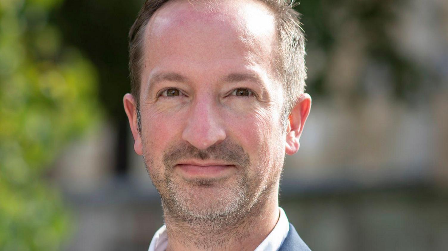 A smiling Michael Solomon-Williams with receding short brown hair and stubble, wearing a white collared shirt and a grey jacket