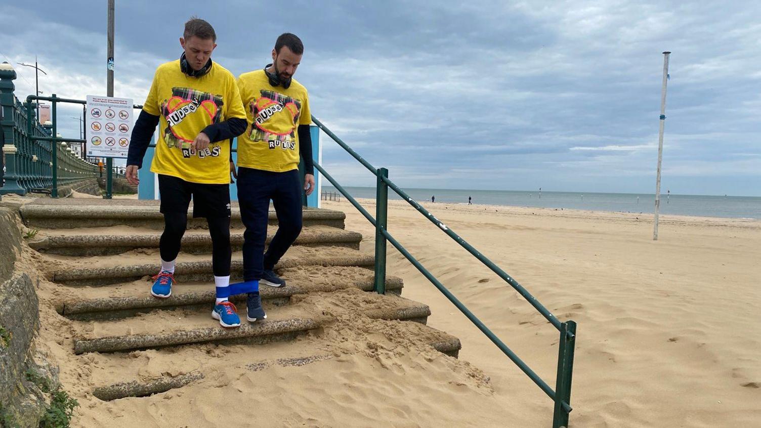 Two men in yellow shirts and black trousers are tethered around their legs and staggering down a flight of stairs at a beach.