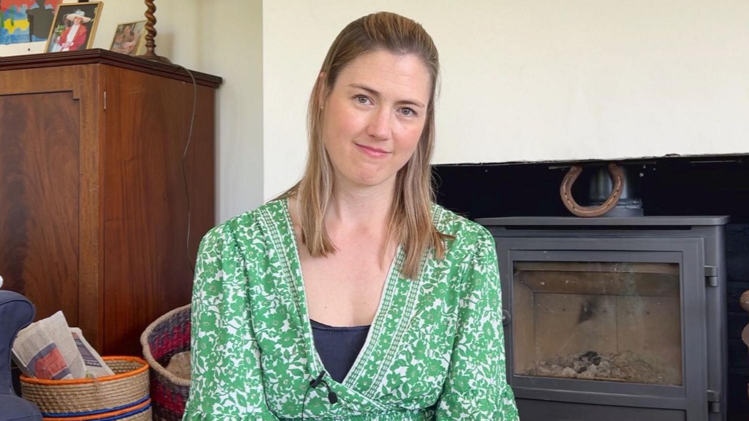A woman with shoulder length brunette hair. She is sitting in a lounge in front of a fire. She is wearing a green floral dress and smiling.