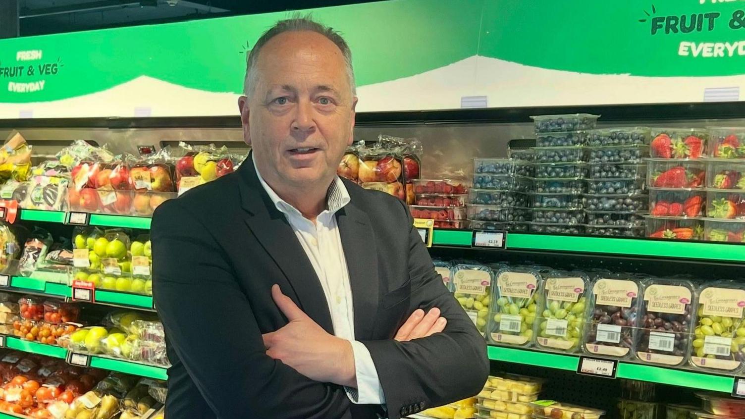 Peter McBride standing in front of a fruit and veg aisle in one of his Spar stores. 
He has short, greying hair and is wearing a dark grey suit and an open-neck white shirt. 