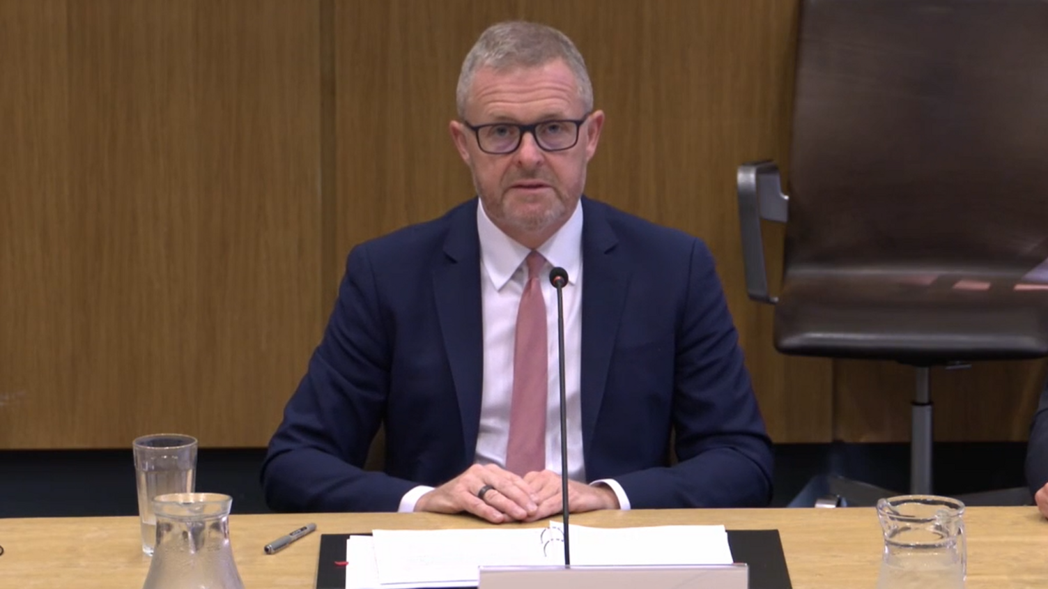 Jeremy Miles sat at a wooden desk, wearing a tie, shirt and a blue suit. A glass of water and two jugs are in front of him, alongside a binder with papers. A chair is behind him.