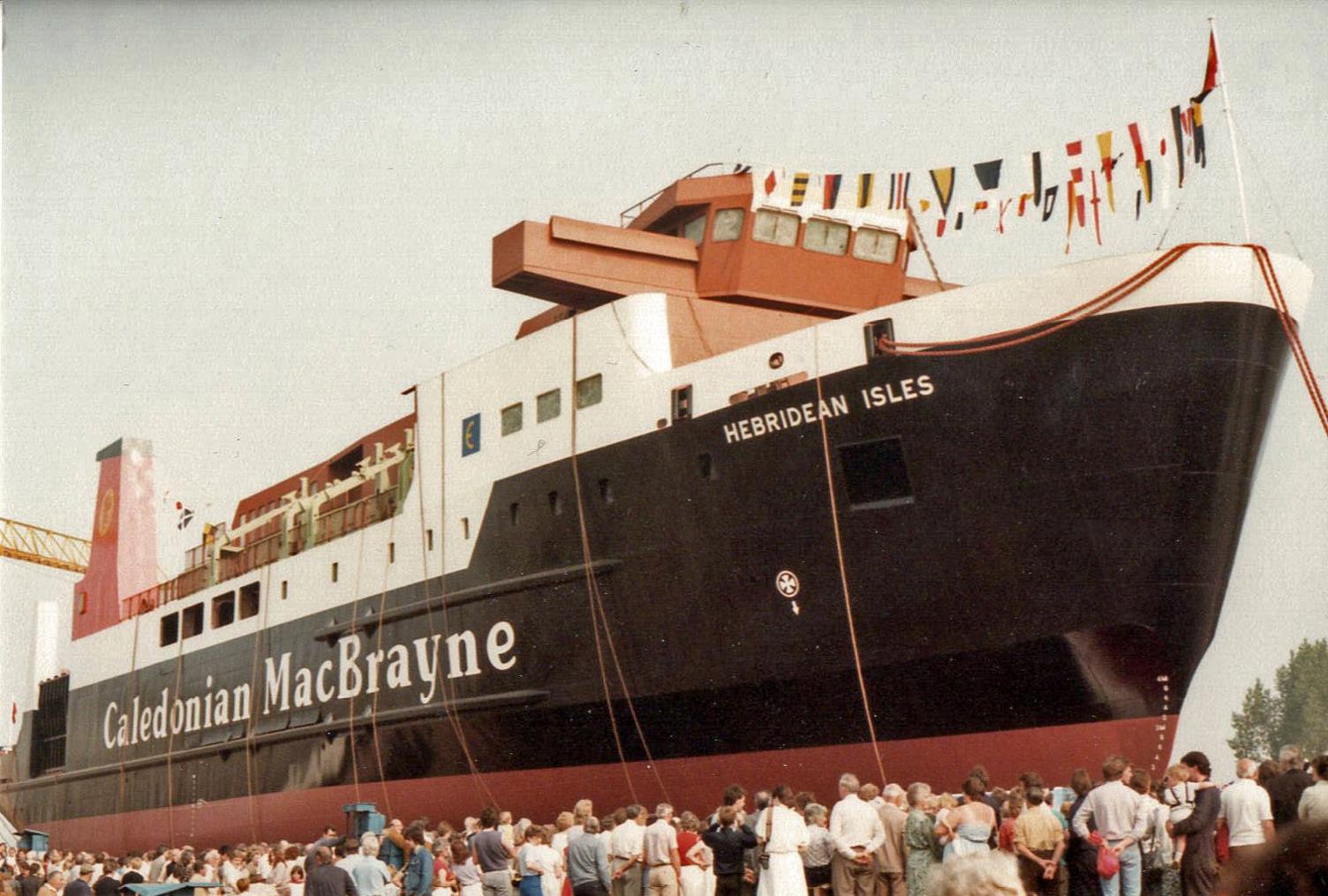 Crowds gather in front of a large black and white ship with a red funnel as it is about to be launched