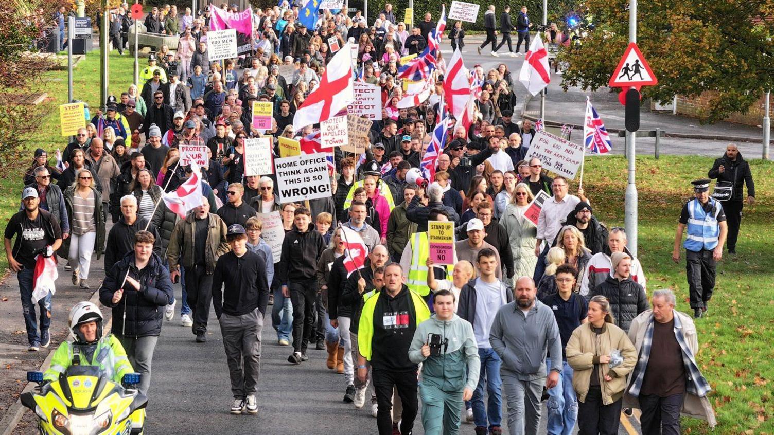 Hundreds of people holding St George flags marching along a residential road.