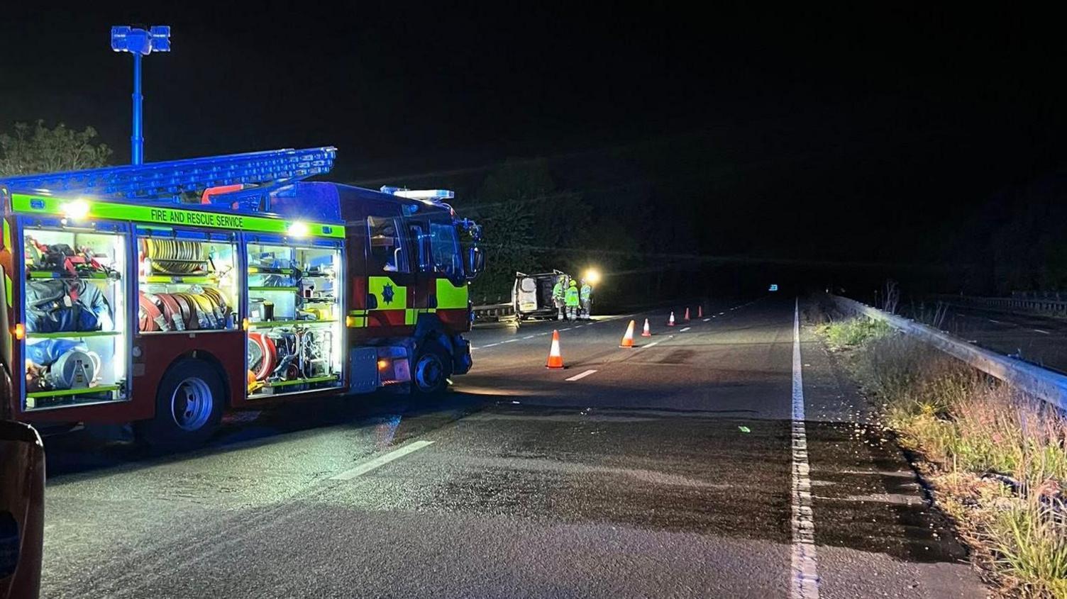 A fire engine, lit up from the inside, parked on the slip road of a dual carriageway. The van is in the distance.