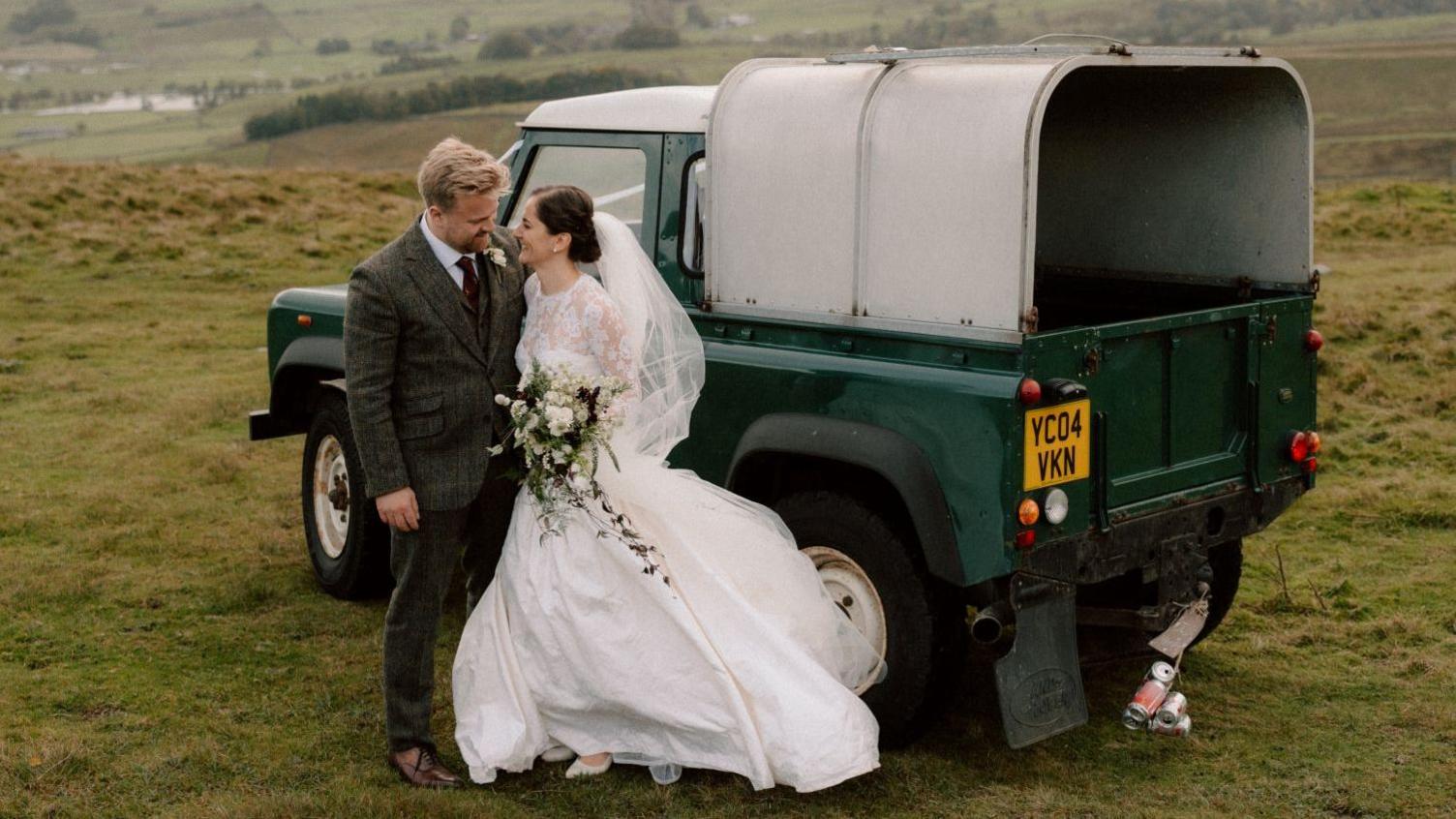 A newly-married couple pose in front of a dark green Land Rover Defender that is parked on a grassy hill. The bride wears a floor-length white gown with lace details and a short veil. She holds a bouquet of white and purple flowers. The groom wears a dark-coloured tweed suit and purple tie.