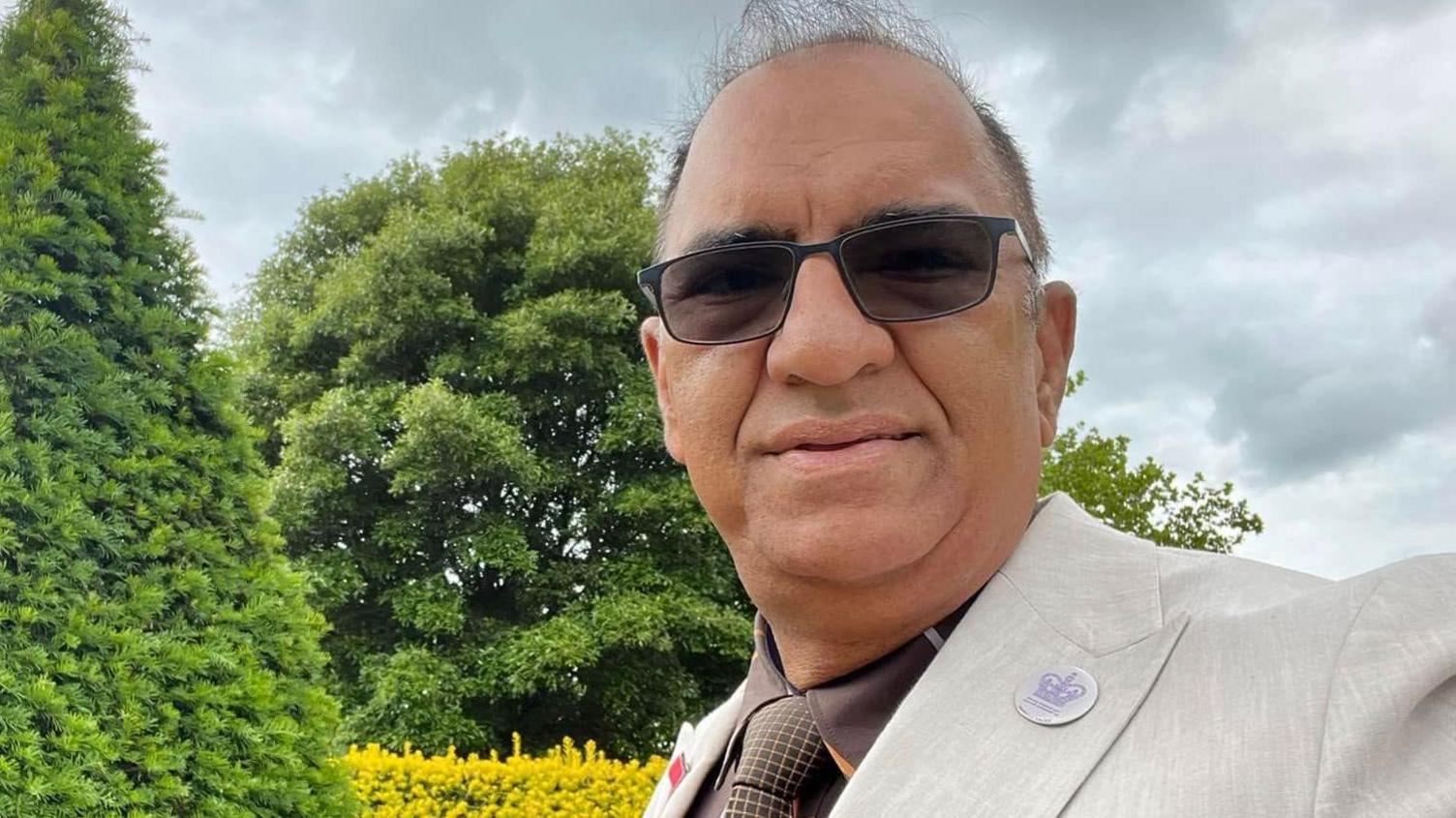 Moez Nathu takes a selfie-style photograph. He is standing outside with green trees behind him. He is wearing dark sunglasses and a beige suit jacket, with a brown shirt and brown tie. He has a button on his lapel. 