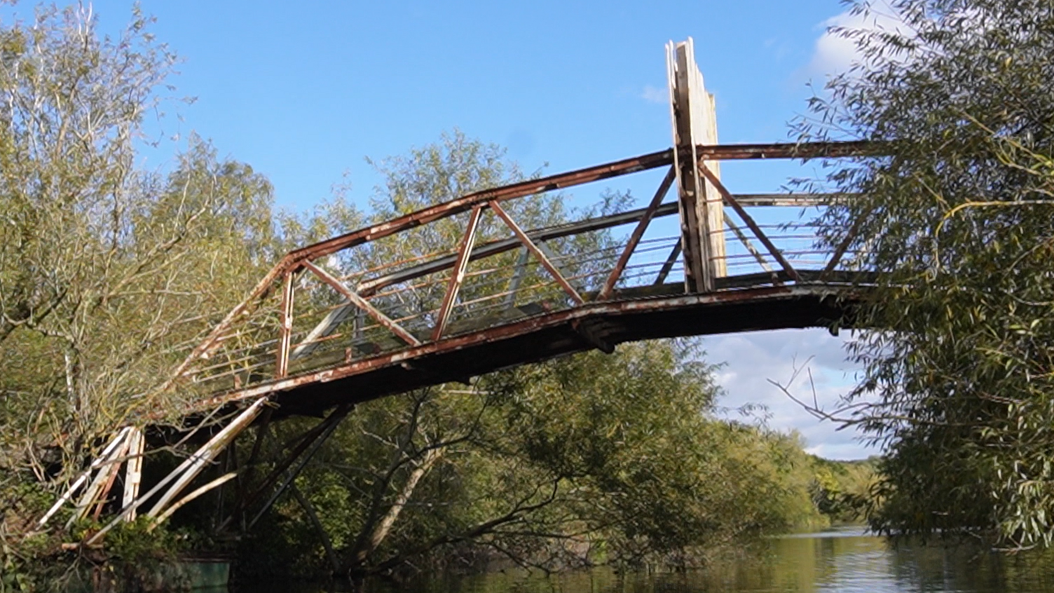 The Works bridge at Attenborough Nature Reserve from the view of the river below, which shows a rusted structure and a wooden board across the middle of the bridge to stop people from using it.