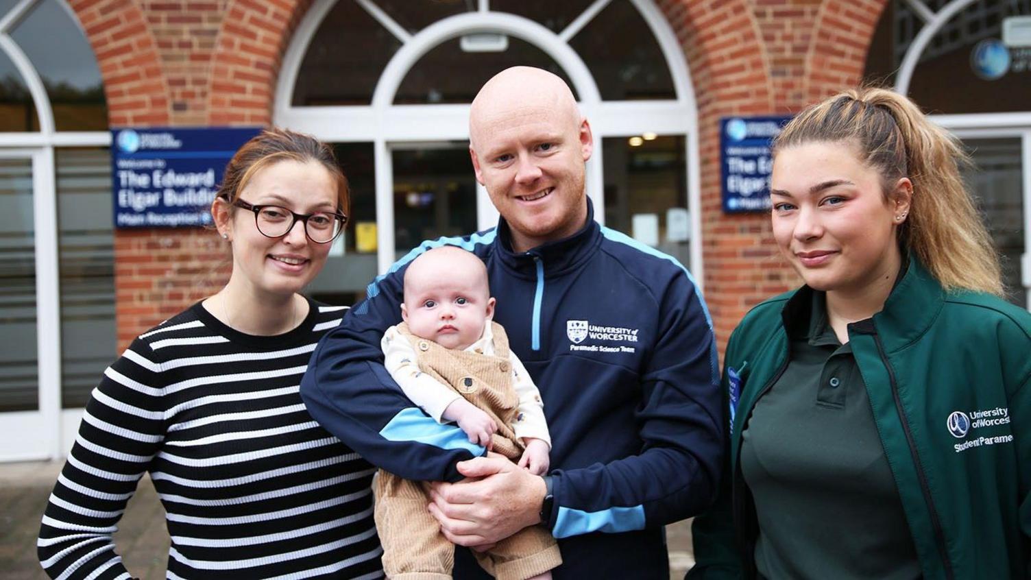 Two women standing either side of a man who is holding a baby. On the left is the baby's mother, who has ginger hair tied back and is wearing glasses and a black and white striped top. The man, the baby's father, is bald with ginger facial hair and is wearing a navy top that says "university of Worcester" on it. on the right is a student paramedic who has light brown hair tied in a ponytail. she is wearing a teal t-shirt and jacket, with the university of Worcester logo on it