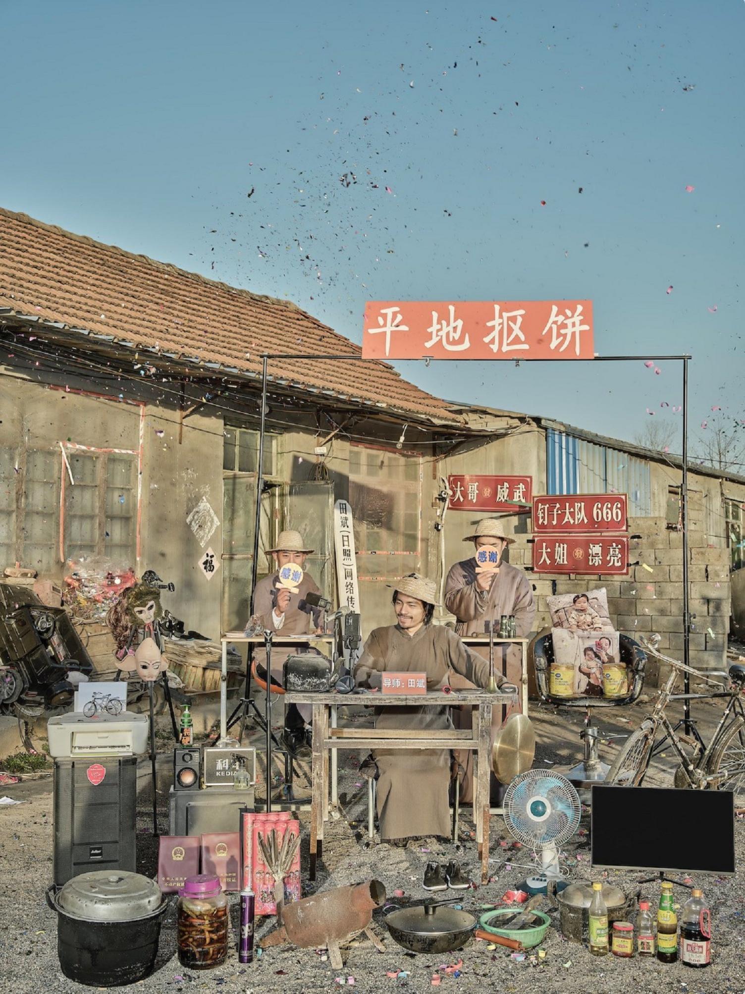 Man dressed in a straw hat and a grey robe sits at wooden bench, flanked with items like bicycles, aluminium tins and rubber masks.