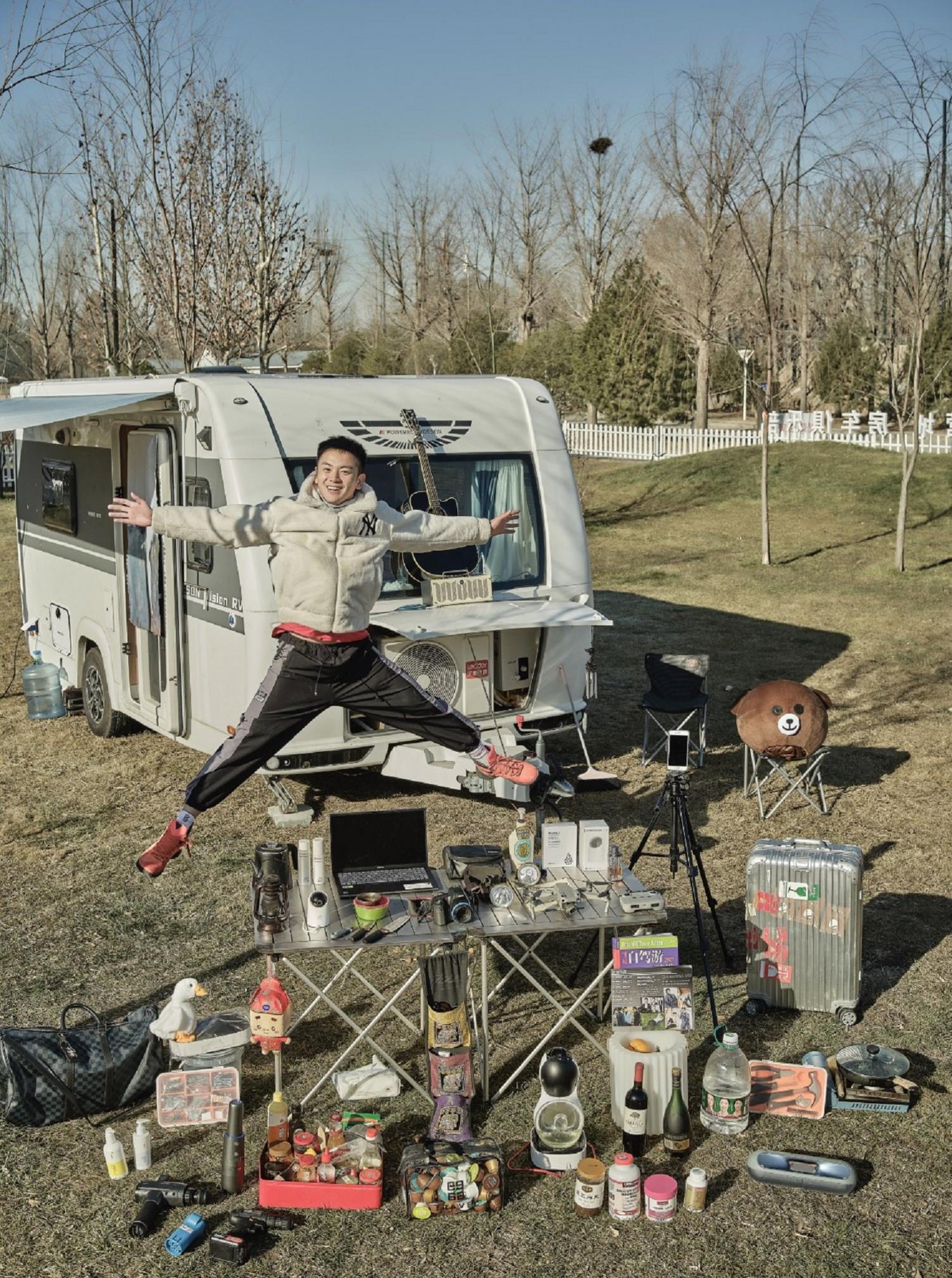 Man jumps in the air in front of a caravan. In front of him on a foldable table are his cooking utensils, foodstuffs and other essential items needed for living in a caravan.