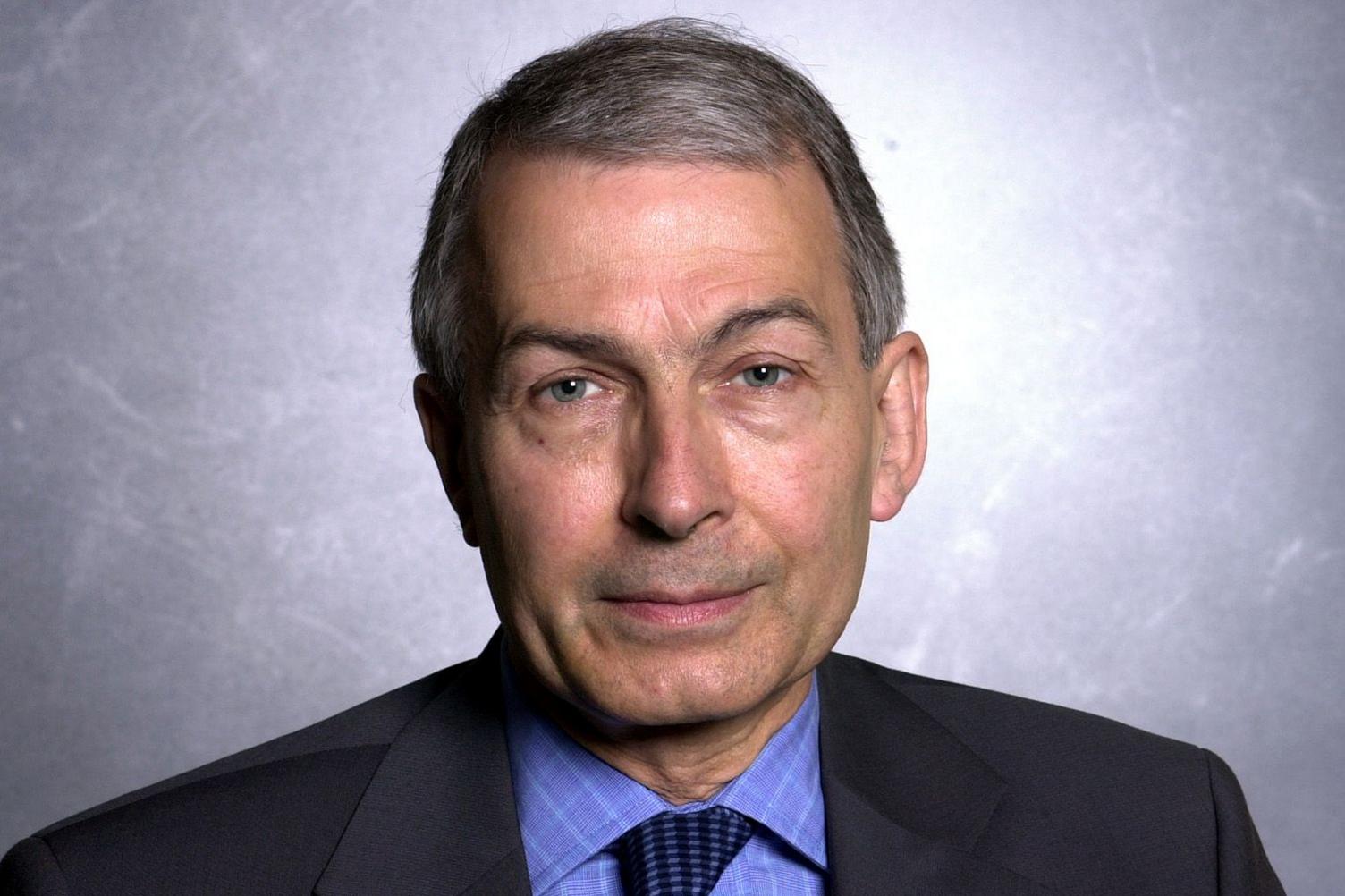 Head and shoulders shot of Frank Field. He has very neatly cut and combed grey hair, and looks into the camera. He is photographed against an anonymous background and is wearing a dark suit with blue shirt and tie.