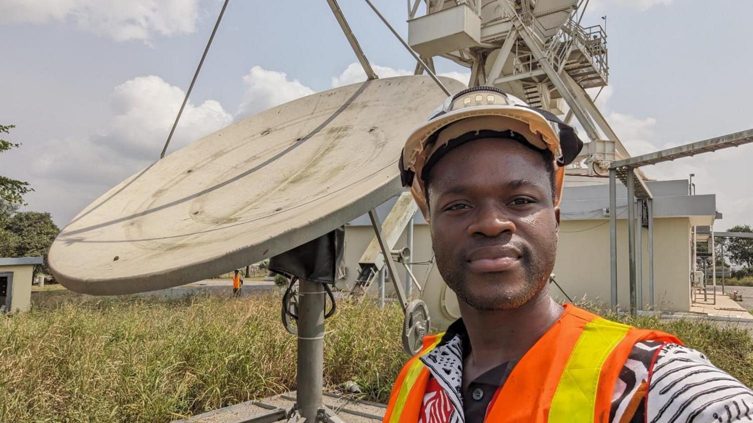 Kwaku Sumah, stands in front of a radio dish wearing a hi-visibility vest.