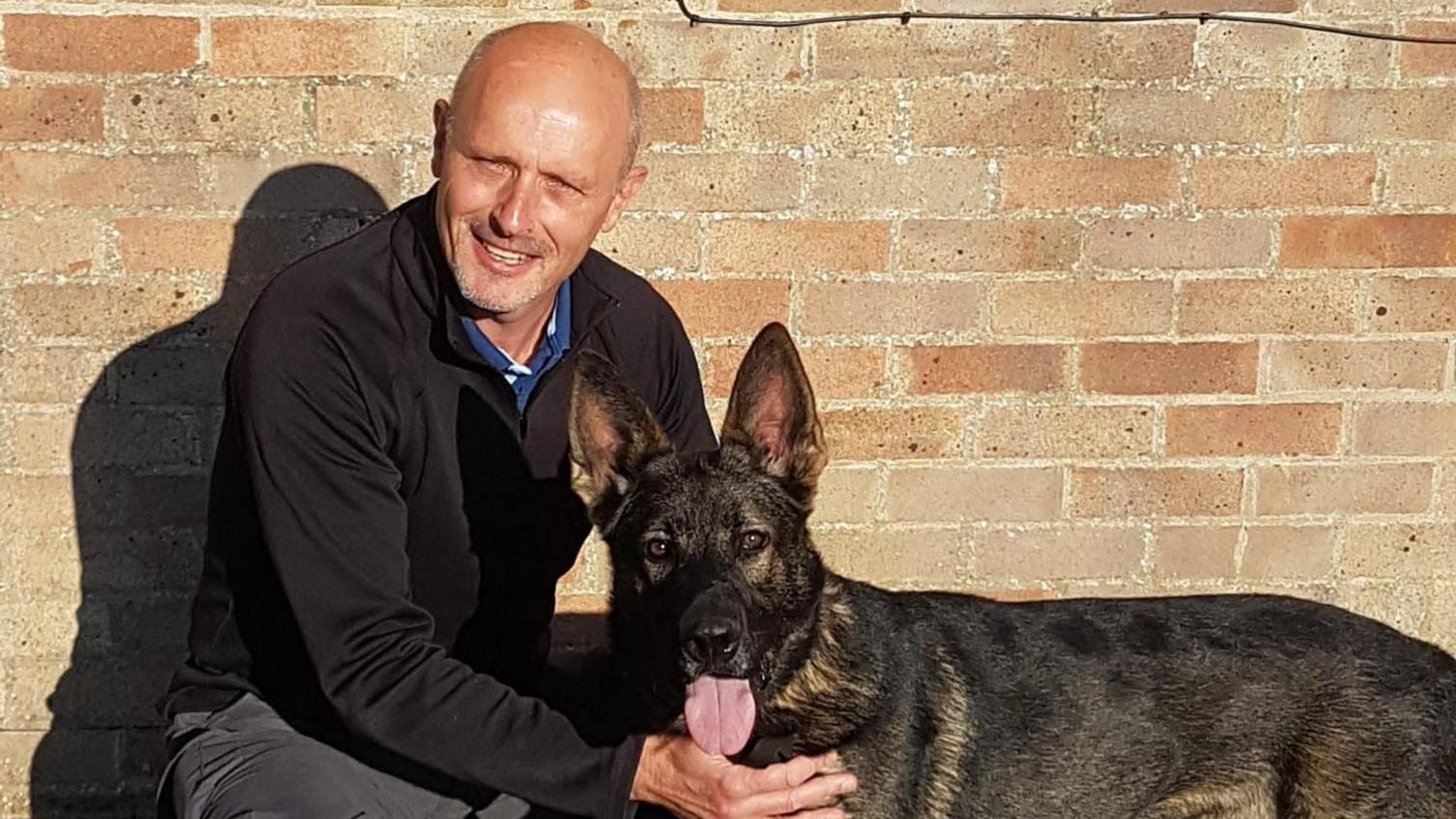 Ian Partington kneeling, holding a dark brown German Shepherd, which has its tongue out. Ian, who is bald, is wearing all black and smiling. They are in front of a brick wall.