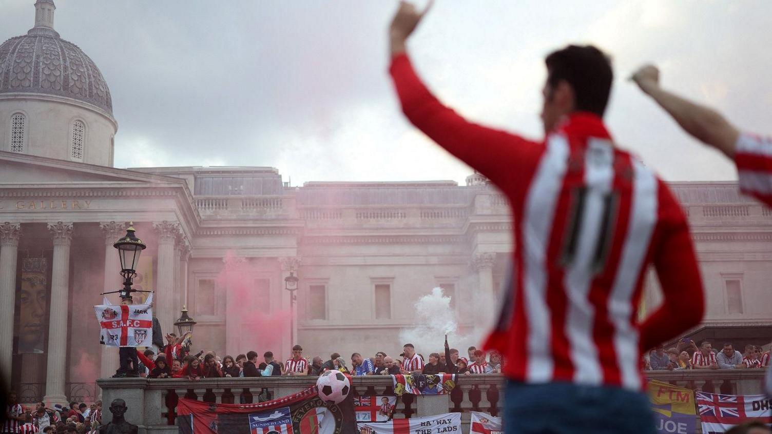 A male Sunderland fan stands with his arm in the air and he wears a red and white top. He is out of focus. Hundreds of fans are in the distance at Trafalgar Square.