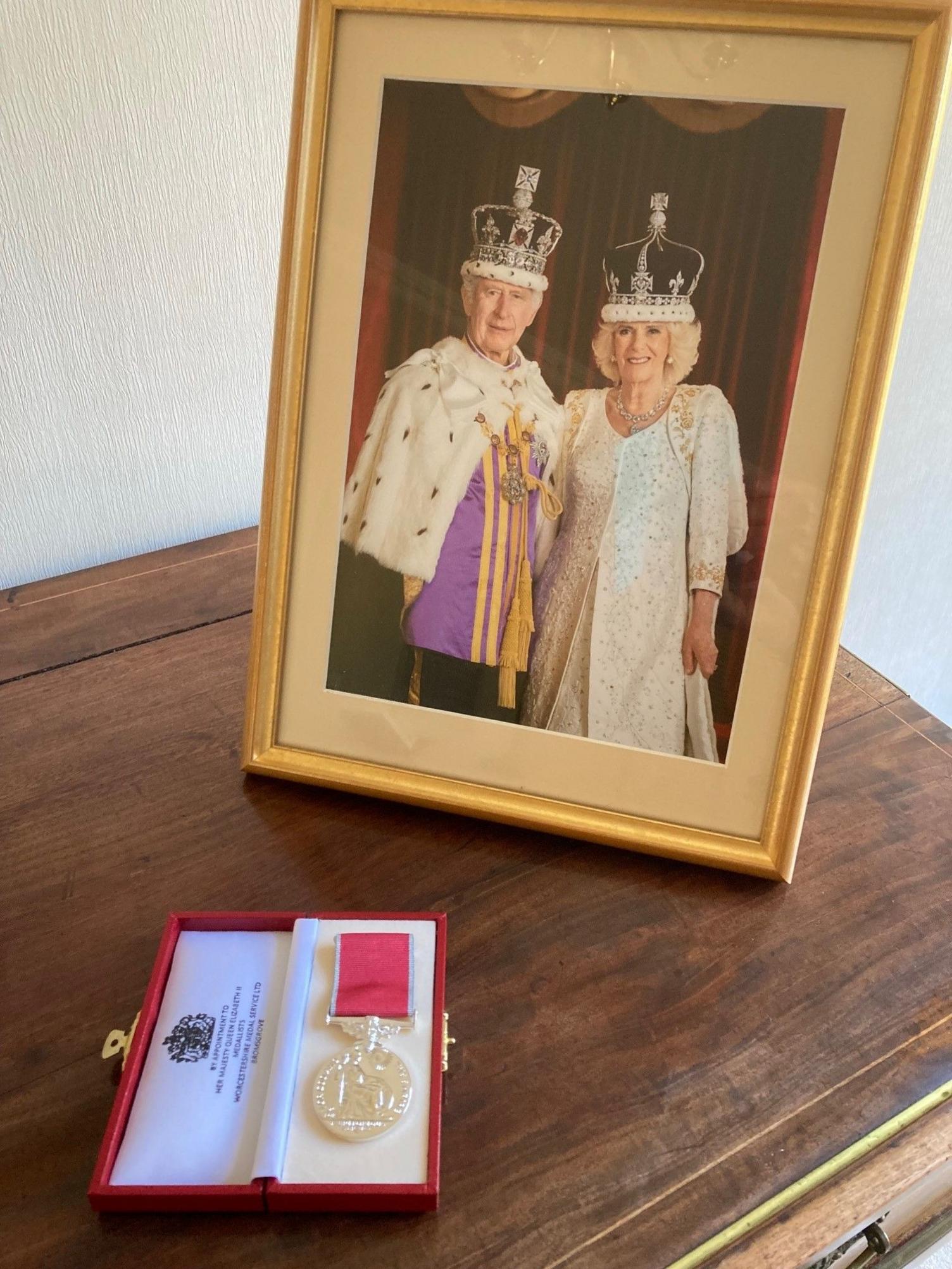 A medal on a side table with a photo of King Charles III and Queen Camilla behind.