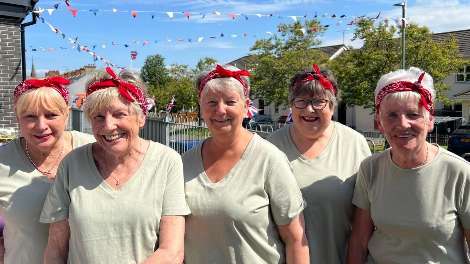 Helen Walker, Jeanette Warke, Donna Best, Annie Shields, Carol Wright are dressed as WW2 'Land Ladies'.
They are wearing red headbands and grey coloured T-shirts.
They are standing in front of a number of flags and bunting.
