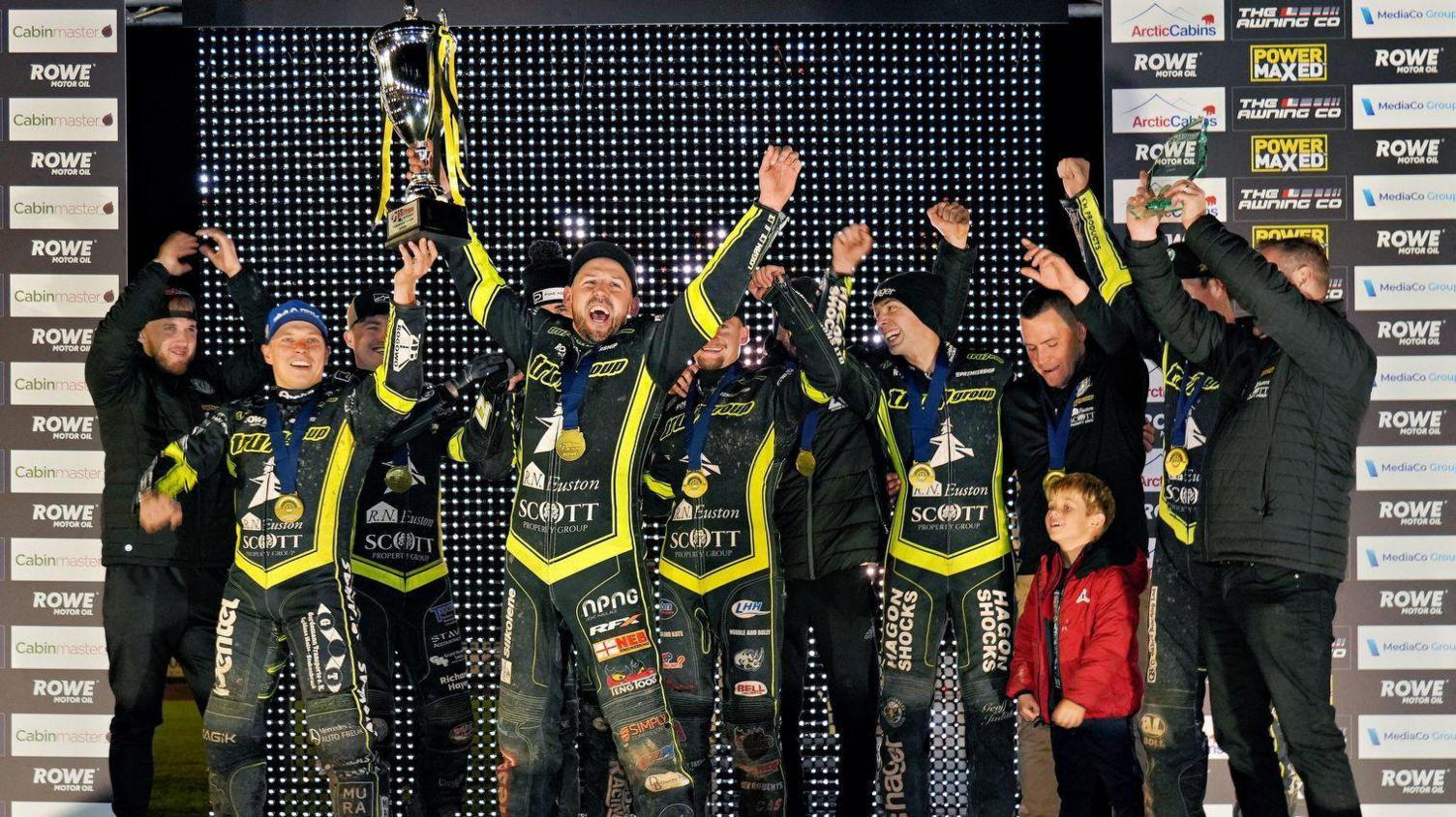 The Ipswich Witches team standing on a stage, cheering with their arms in the air, celebrating. They are all wearing motorcycle leathers and medals around their necks, while one of the riders is holding the trophy aloft.