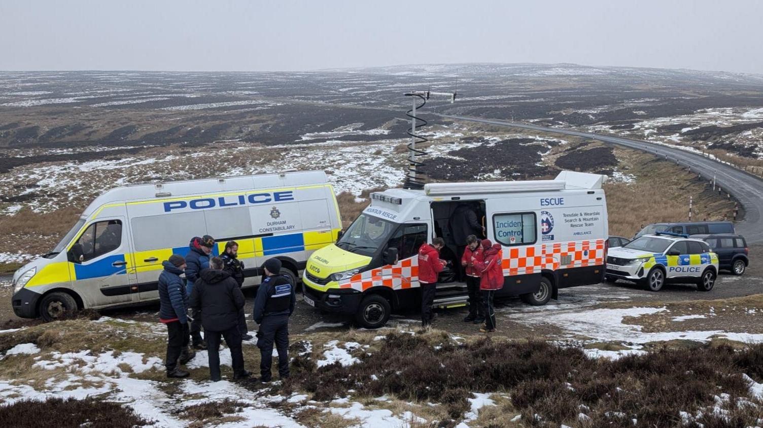 A  police van parked alongside a mountain rescue van. Mountain rescuers can be seen in front of the vans in red jackets with police officers in black standing in a separate group. Moorland can be seen behind the vans which stretches beyond the horizon.