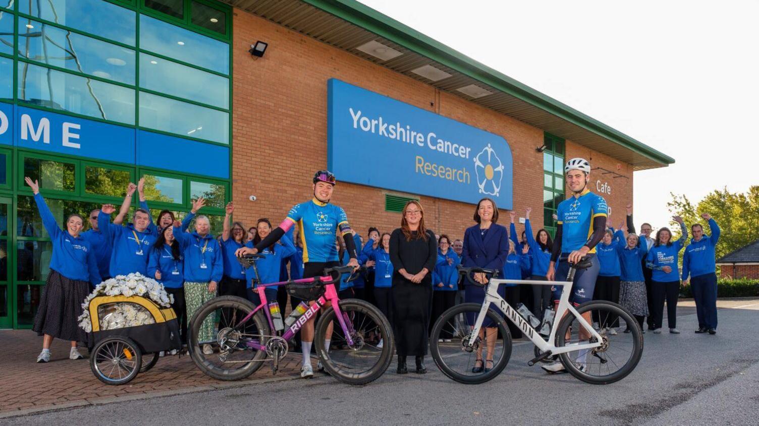 Two men with bicycles stand in front of the Yorkshire Cancer Research building with others cheering and whooping in the background. A trailer full of white roses is attached to one of the bikes.