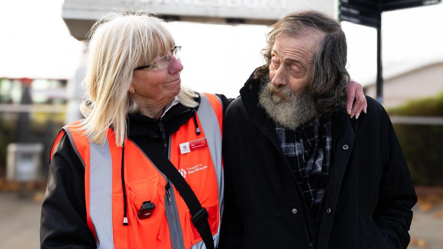 A lady with blonde hair and wearing glasses, with her arm around an older man with grey hair and a beard. They are looking at each other. 