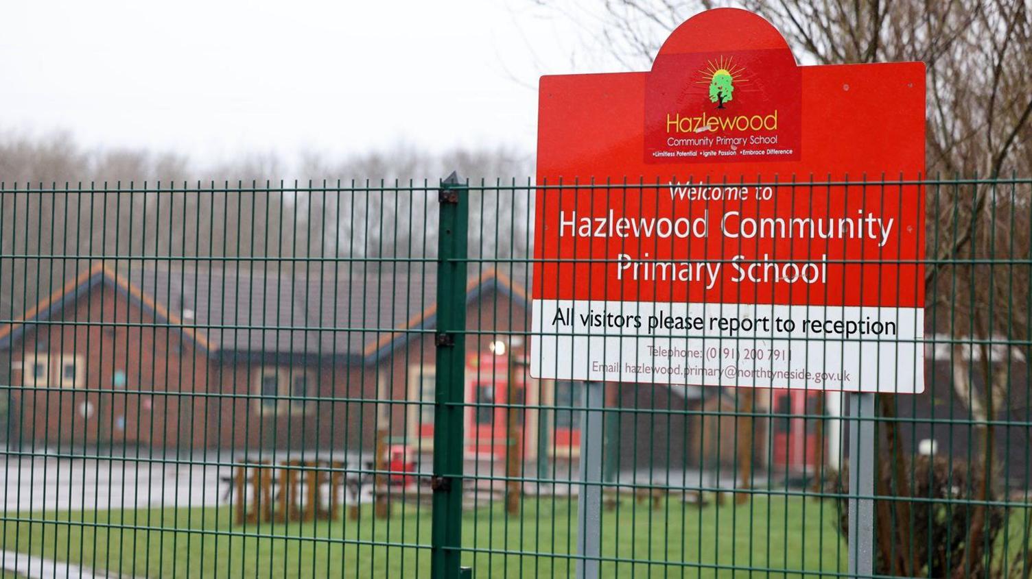 The outside of Hazlewood Community Primary School which is a red brick, sing-storey building. There is a red sign in front of it with the school name and logo, which is a green tree. There is a green fence in front of it.