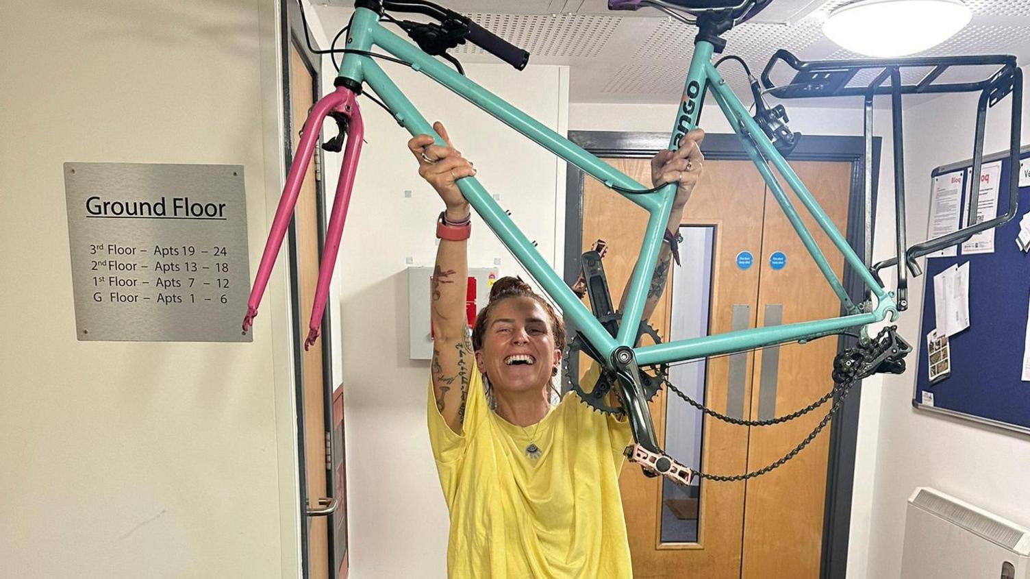 Fiona is wearing a yellow T-Shirt and grey jogging bottoms and pink shoes. She is holding her bike frame about her head. It is missing two wheels. 