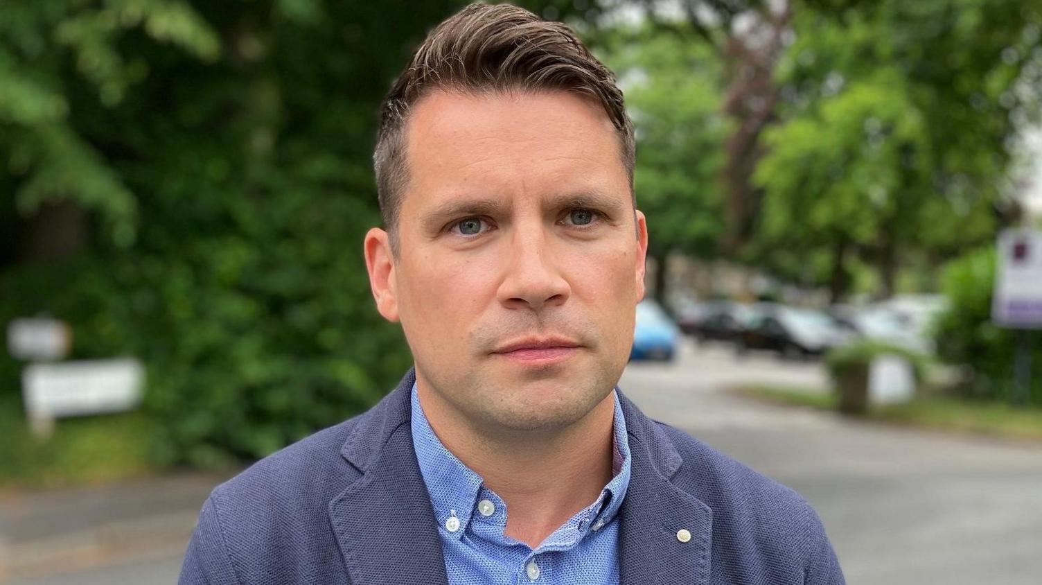 A man with short brown hair wearing a blue shirt and navy jacket stands in a carpark