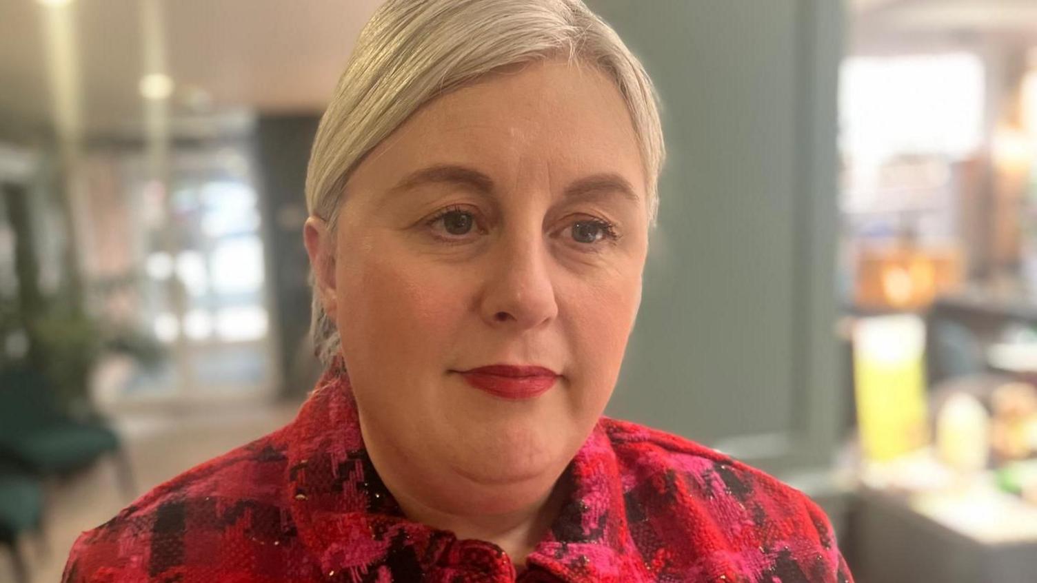 The head and shoulders of a woman in a bold red jacket with her blonde hair tied back stood inside