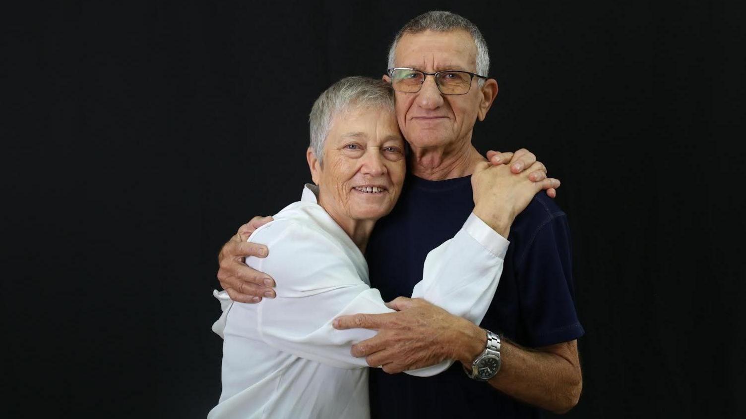 Bilha and Yakov Inon posing for a photograph against a black background. Bilha is wearing a white shirt and her husband is wearing a black t-shirt and he has glasses on.