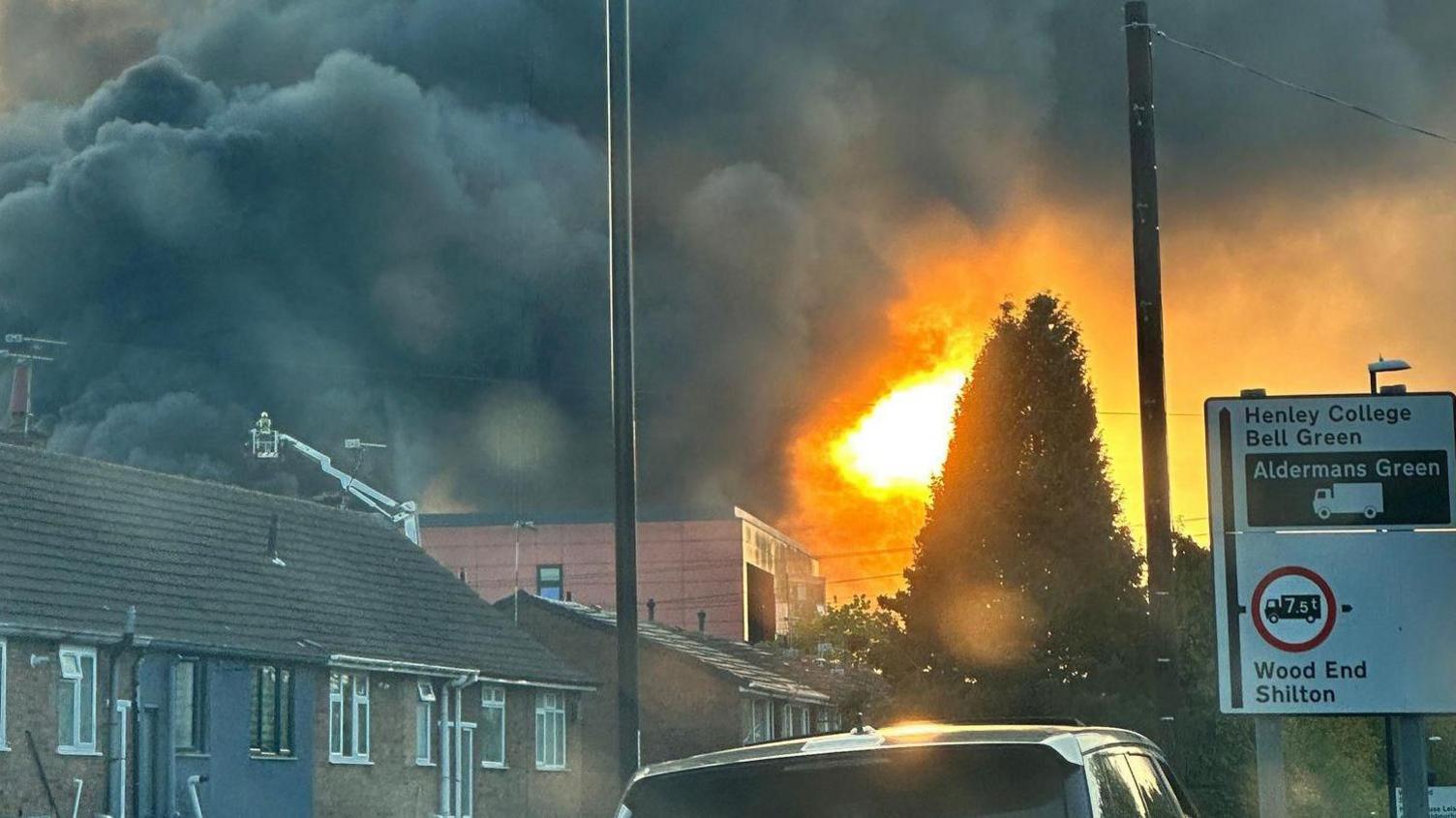 A residential area, with a black car In the foreground. Black smoke and orange flames can be seen over houses