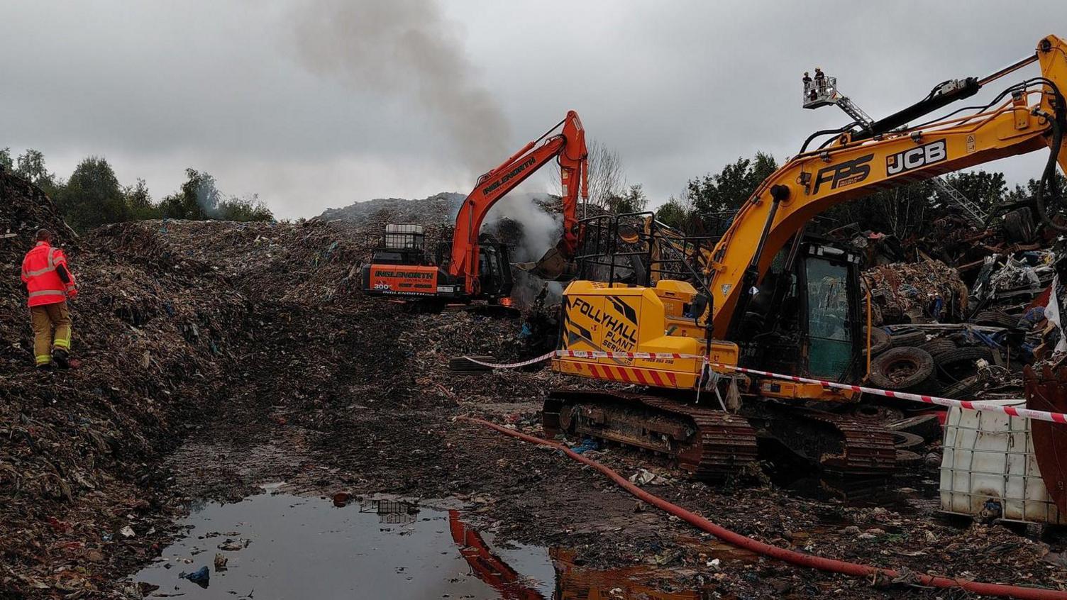 Excavators are parked in the middle of a large waste site. A man in a high vis jacket can be seen walking through it.