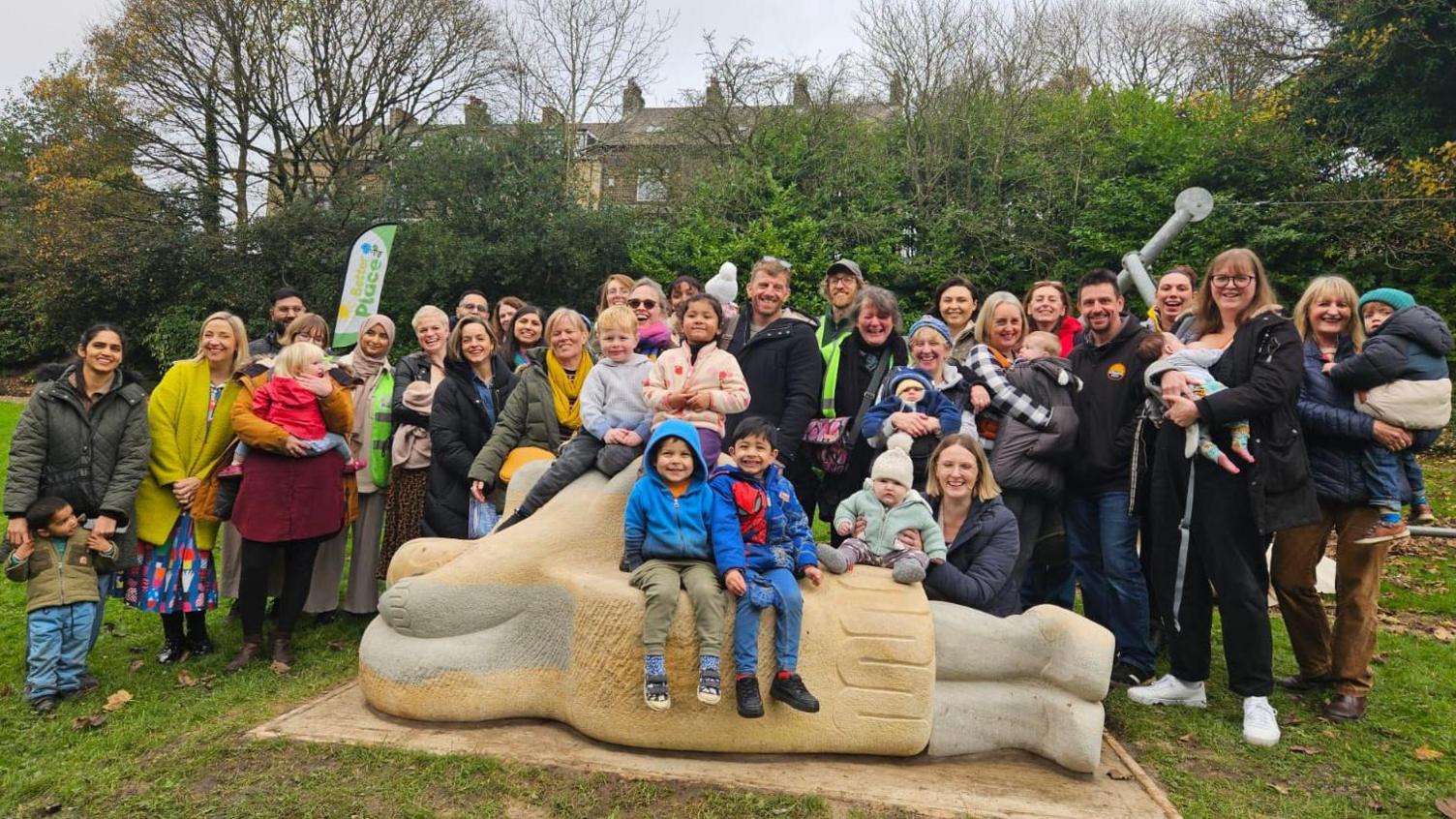 A large group of people - mums, dads and young children - stand with a large stone sculpture which depicts a woman lying on her side breastfeeding. Several children sit on the sculpture's 'seat' section.