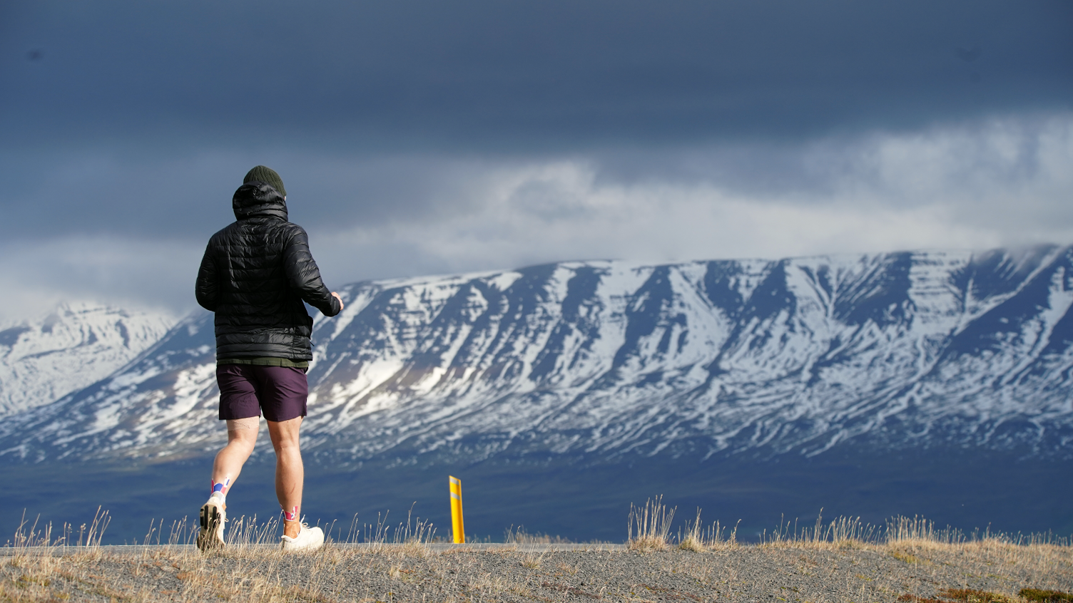 A photo of a man, taken from behind, wearing a black waterproof coat, purple shorts, a dark green woolly hat and white trainers, running along a flat road with a row of snowy mountains in the background.