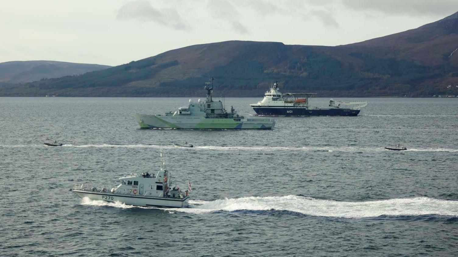 Three of the Rattlers can be seen moving through the water. They are surrounded by three larger vessels guiding them. In the distance is dark green and brown hillside.