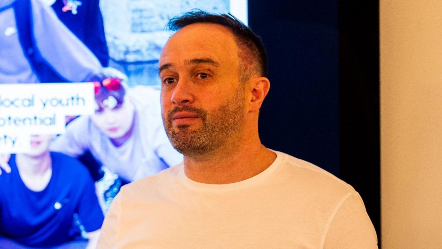 Stephen Dallas has short black hair and beard stubble. He is wearing a white tee-shirt and is standing in front of a display screen showing a group of young people with the words 'Vision' and 'Mission' written on it.