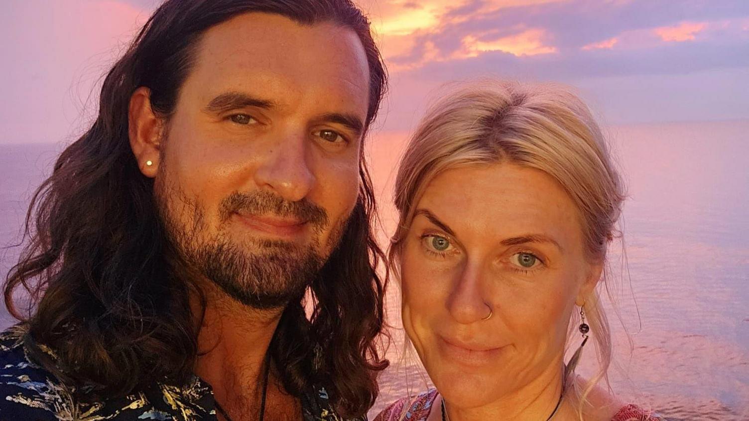 A man with long, curly brown hair an a stud earing poses with a woman with tied back blonde hair and feather earrings. They are standing in front of a sunset on a beach.