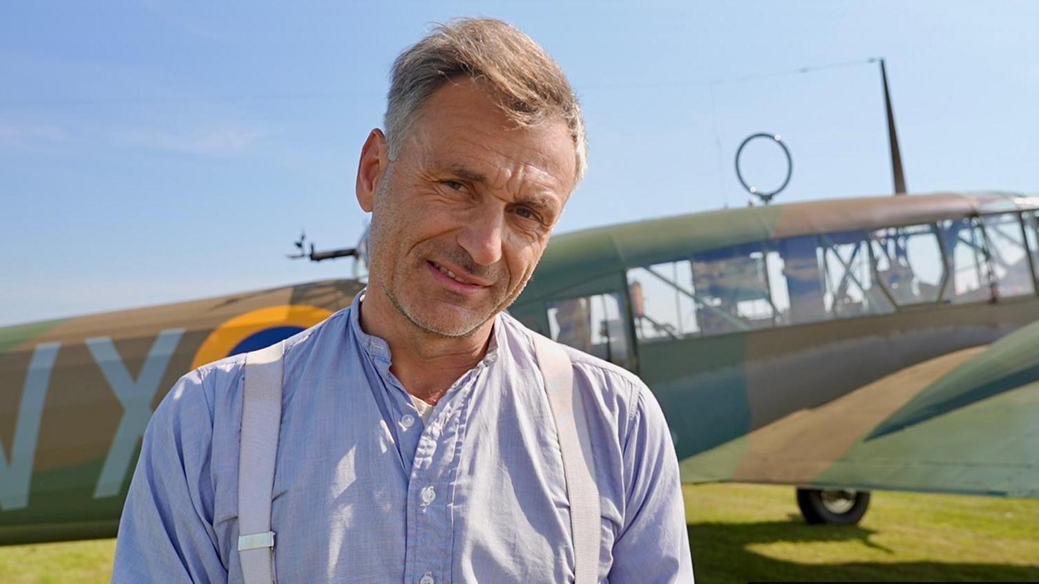 Mr Santus is standing in front of the aircraft on a sunny day at the Duxford airfield. He has grey hair and facial hair and is smiling at the camera with his head tilted to the side. He is wearing a light blue shirt with braces. The Anson plane is behind him.
