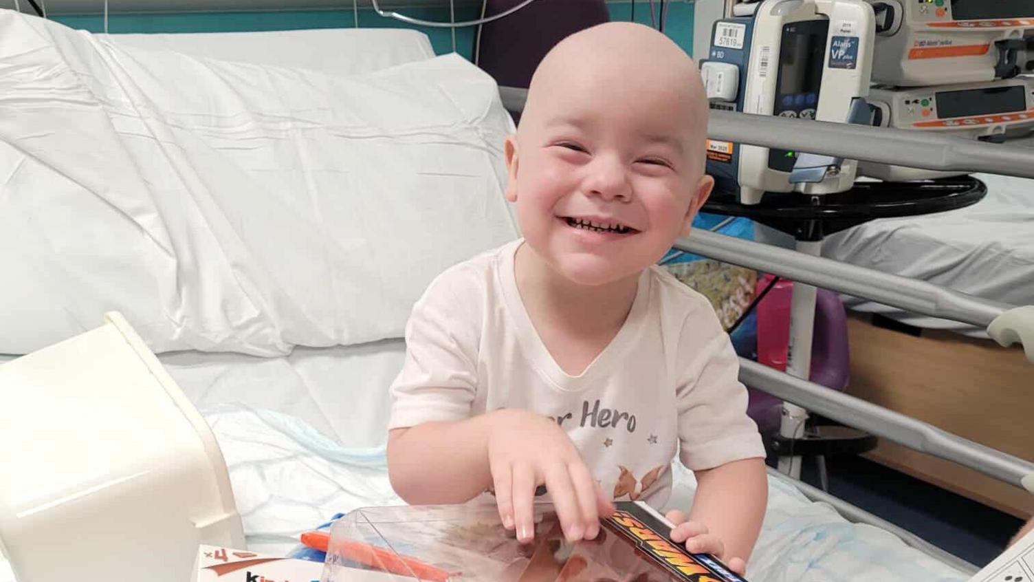 Louie, who has lost his hair and eyebrows, smiles at the camera from a hospital bed. He is opening presents. 
