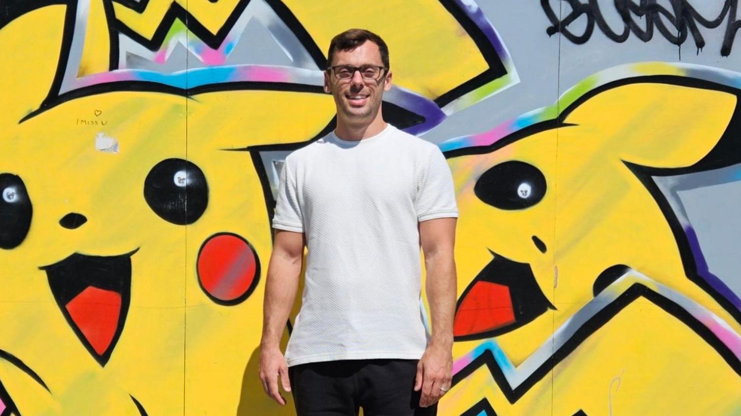 Mr Free is pictured in a white t-shirt smiling to camera while he stands in front of a wall that has been graffitied over with images painting of the Pokémon Pikachu.