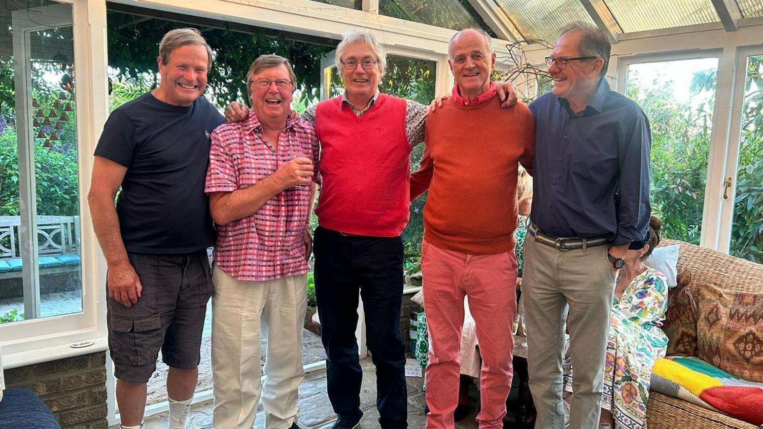 Five men standing with with their arms around each other smiling to the camera, standing in a conservatory with bushes seen in the background.