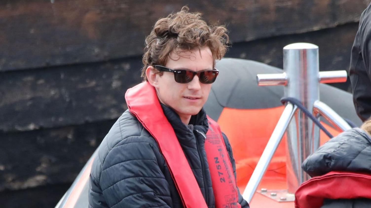 A close up of Tom Holland, wearing sunglasses and a red life jacket over a dark jacket, is sitting in an inflatable boat. He has his brown hair in his trademark tousled style. Behind him is a grey harbour wall.