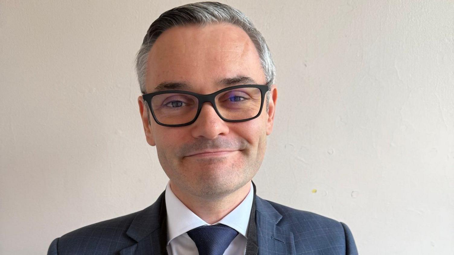 A man with black and grey hair swept back and glasses in a blue checked suit with a light blue shirt and a blue tie