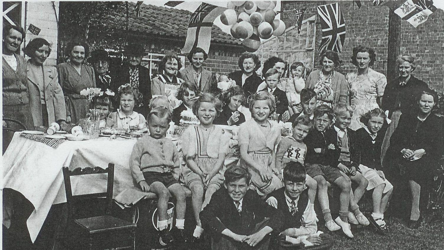 A black and white photo showing a group of people, mainly children, in 1950s clothing, sat around a table smiling at the camera, with union flags hanging in the background.