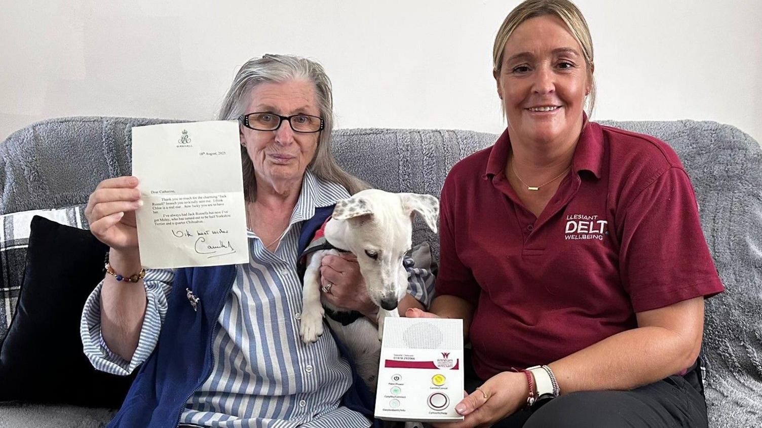 Two women sat on a grey sofa. One with grey hair and glasses, holding up a letter from Queen Camilla in one hand and a Jack Russell dog in the other. The second woman has blonde hair and a dark red polo shirt, with the words Delta Wellbeing sewn in