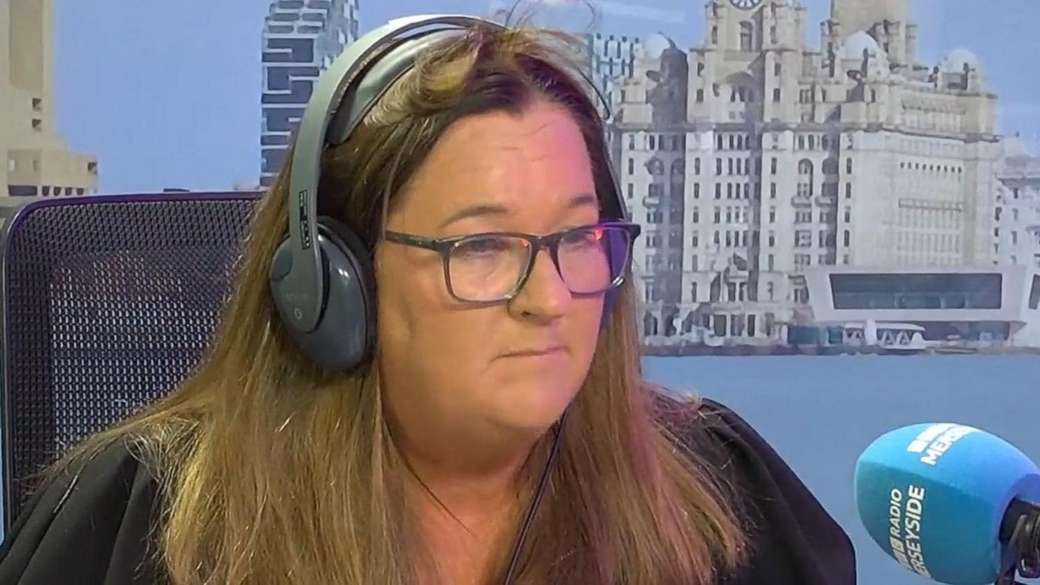 Marion Atkinson has long, light brown hair and is sitting in a BBC Radio Merseyside studio wearing headphones behind a blue microphone. She is wearing a black top.