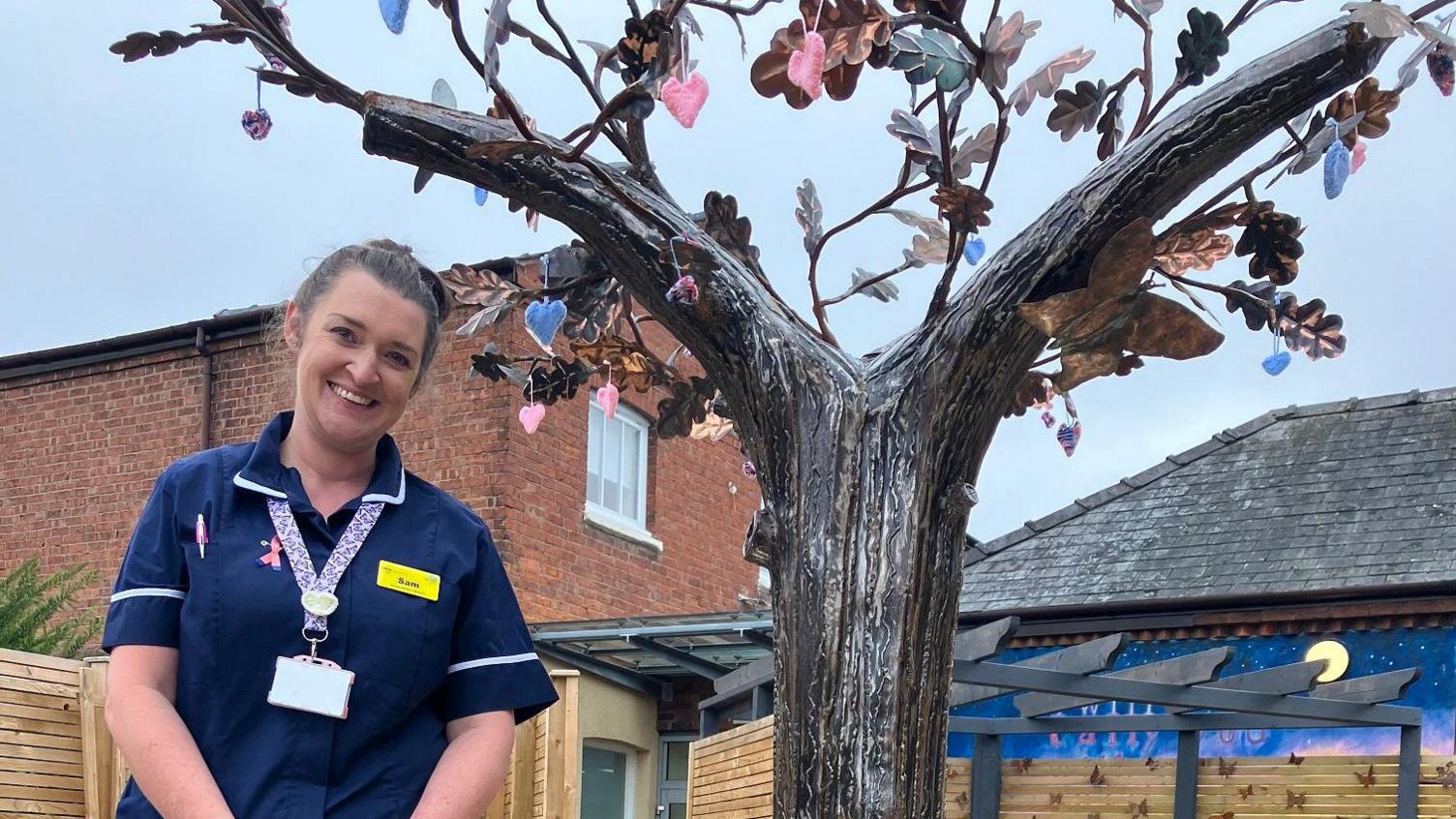 A woman with brown hair tied back, smiling at the camera. She is wearing dark blue scrubs with a purple and white lanyard and a yellow name badge. She is standing in a garden that has a mural on a wall in the background. Next to her is a tree sculpture with gold/brown leaves on it. Hanging from the branches are small blue and pink hearts.