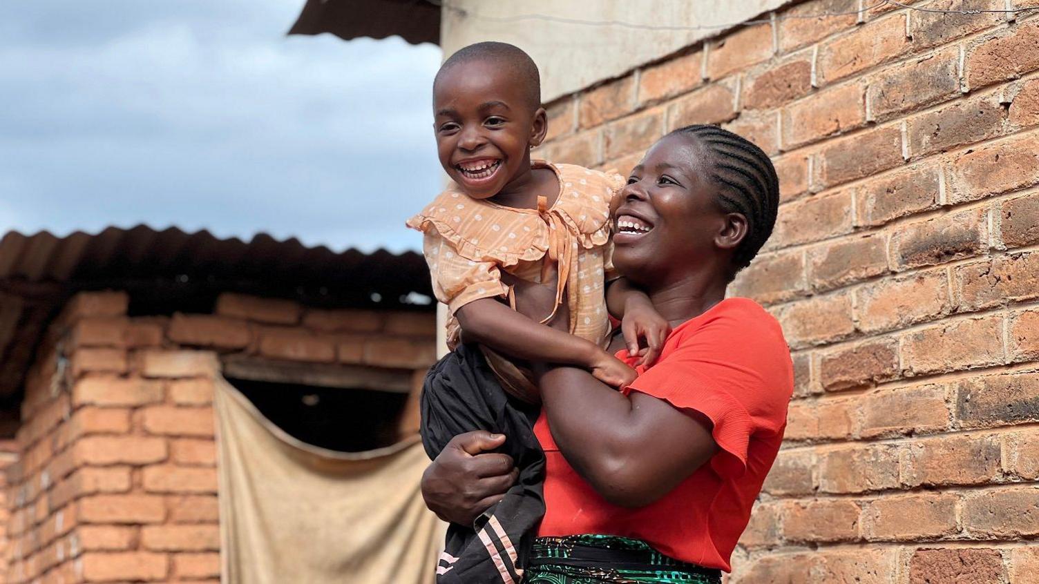 A young girl is being cuddled by a woman in a red top. They are outside and both smiling.