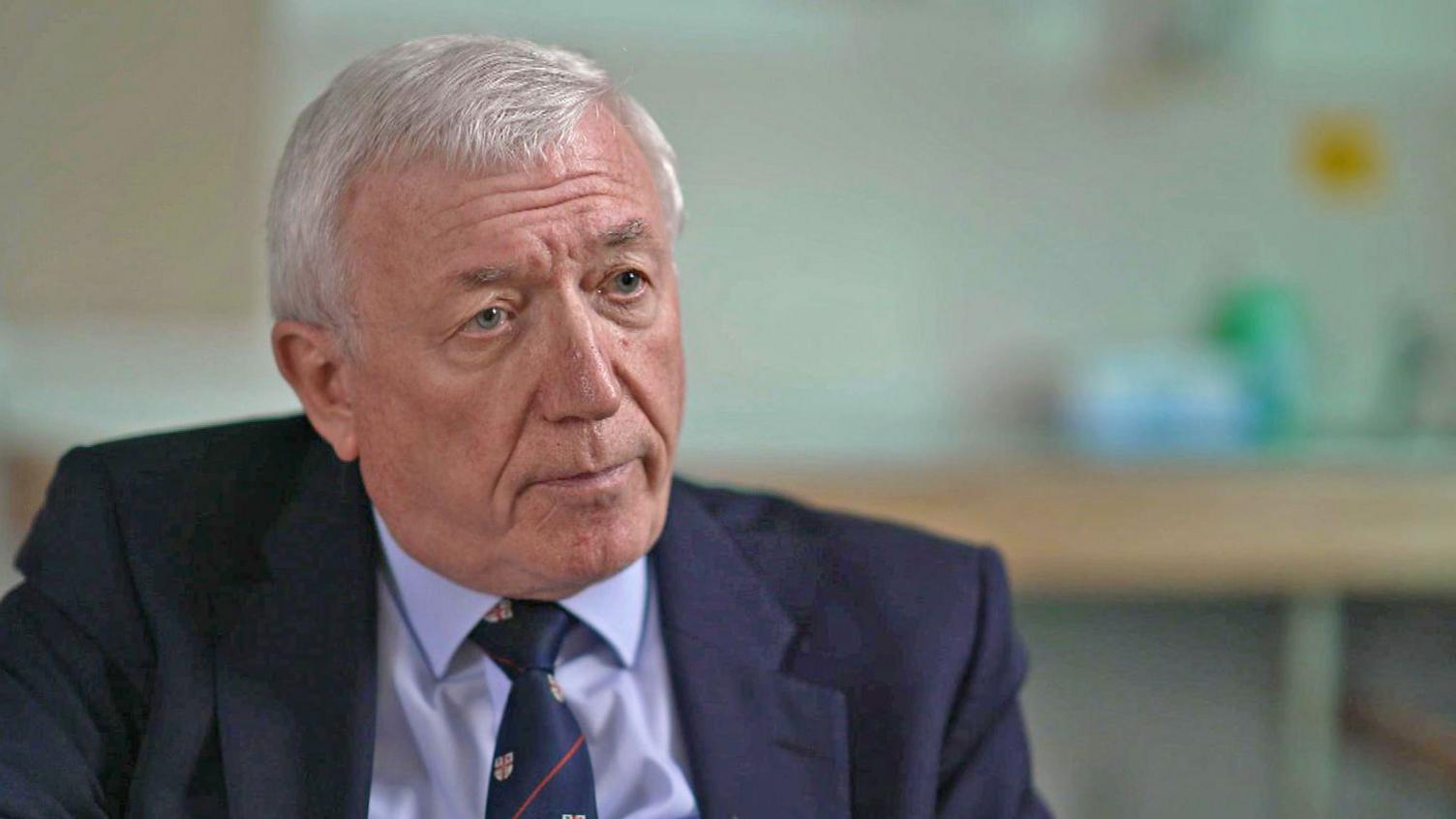 Head and shoulders shot of retired consultant neonatologist, Mike Hall - a man with straight grey hair, wearing a dark blue suit jacket, and light blue short and a navy blue tie.