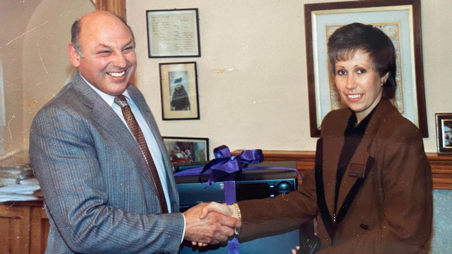 Bob Williams on the left, wearing a grey suit, shaking hands with a woman, presumably the shop manager on the right, who is wearing a brown suit. 