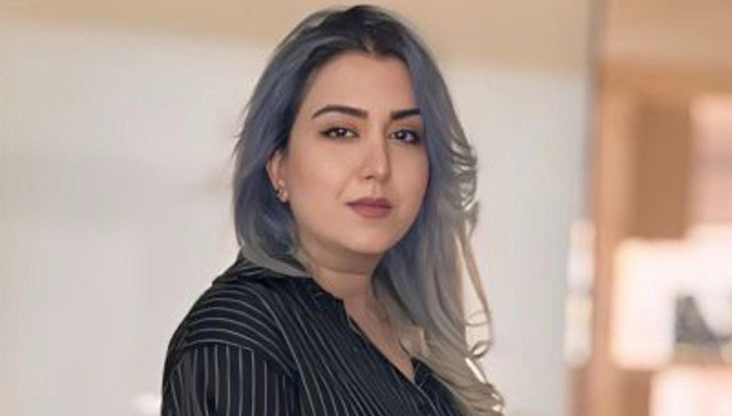 A side profile of Mana with pale purple coloured hair and a pinstriped black top. She is looking at the camera and the background is out of focus.